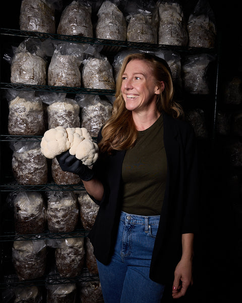 Rachel the founder of MYCOfactor holding white lion's mane mushrooms in front of shelves with mushroom bags in a farm